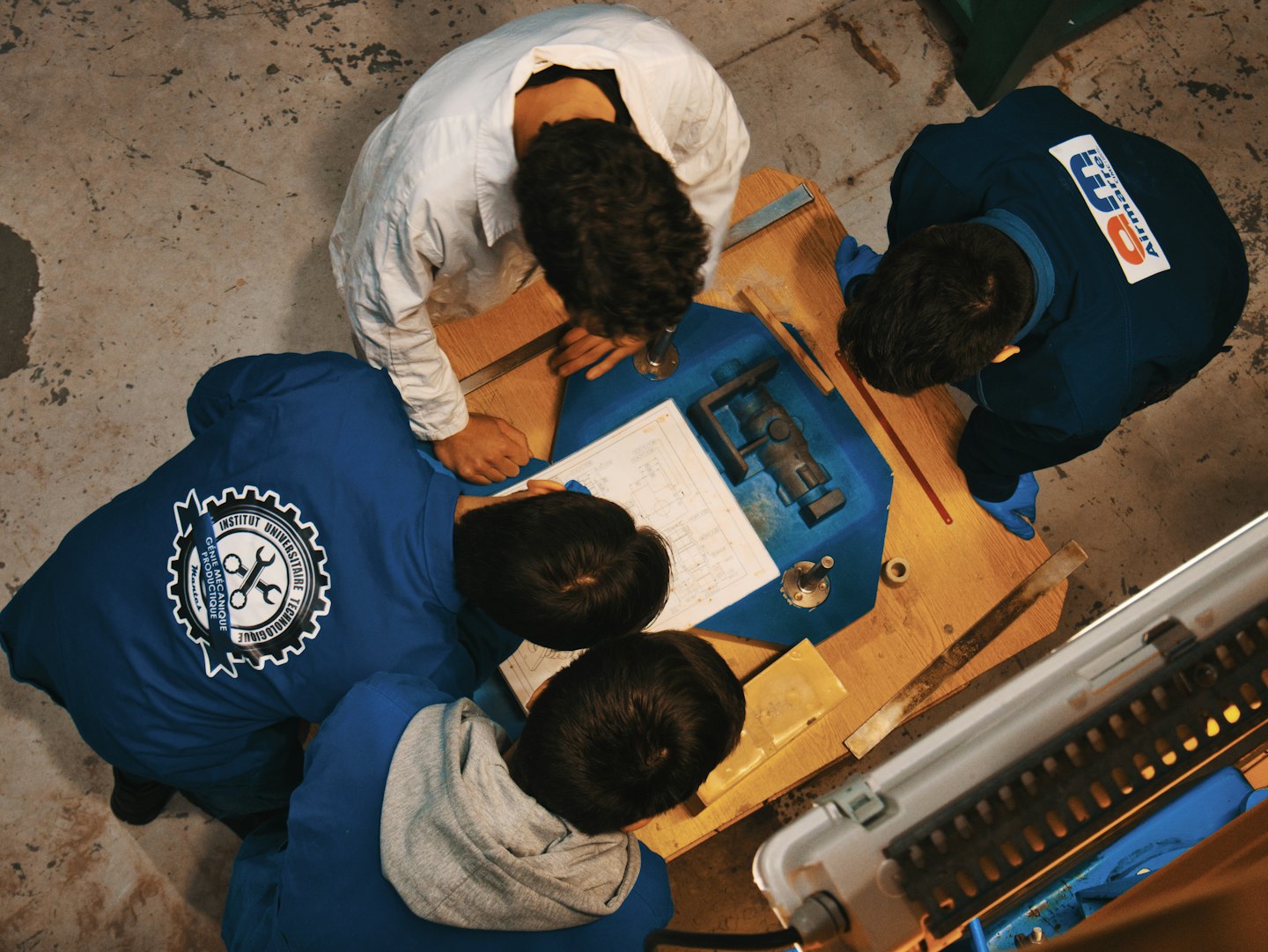 Four people in work uniforms examine blueprints on a workbench.